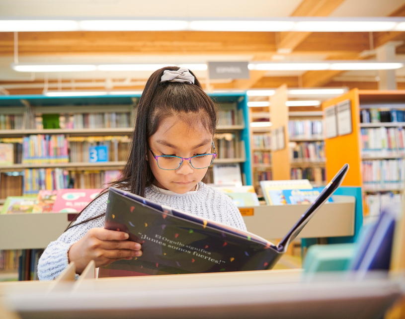 Young girl wearing glasses reading a picture book in the library