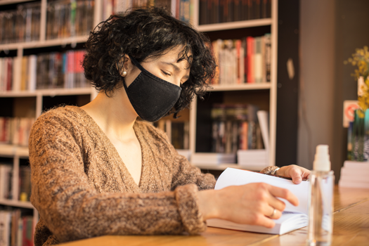 Woman in mask looking at book in library, hand sanitizer on table