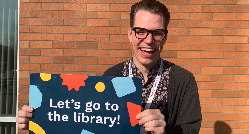 Man with glasses stands outside holding a giant library card for kids