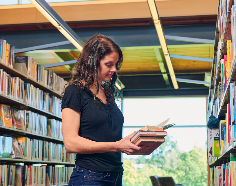 Woman with brown hair wearing blue t-shirt reading book in library